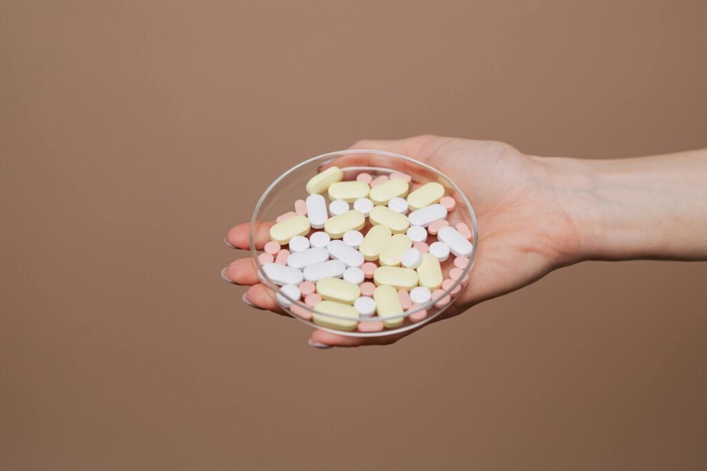 Close-up of a hand holding a petri dish filled with various tablets and capsules on a neutral background.